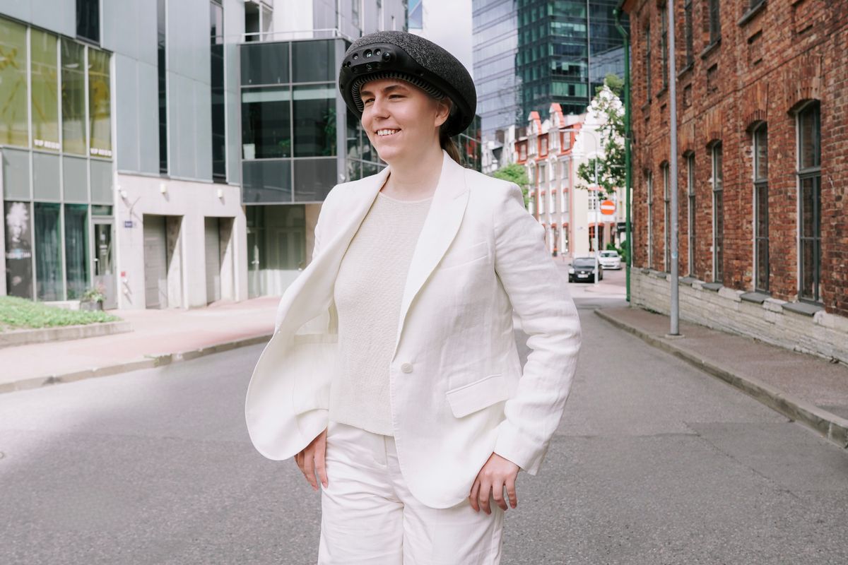 A person in a white suit wearing the 7Sense device on a city street, with modern buildings and a brick wall behind them.